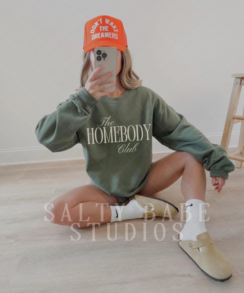 May include: An olive green sweatshirt with "The Homebody Club" printed on the front. The image also shows an orange cap with the text "Don't Wake The Dreamers". The person is wearing tan clogs and white socks.