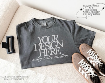 May include: A charcoal gray short-sleeve t-shirt featuring the white text YOUR DESIGN HERE and salty babe studios. The shirt is displayed with a black hair claw clip and a pair of brown and white sneakers on a textured white surface.