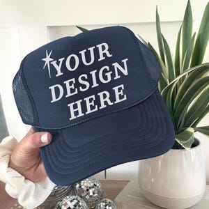 Könnte beinhalten: Eine marineblaue Trucker-Cap mit Netzrückseite und festem Vorderteil. Auf der Vorderseite befindet sich der weiße Schriftzug "YOUR DESIGN HERE" zusammen mit einer kleinen vierzackigen Sterngrafik und einem gebogenen Schirm.