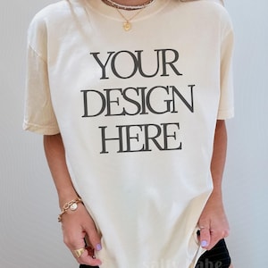 May include: Cream-colored t-shirt with the text "YOUR DESIGN HERE" in black block letters. The shirt is short-sleeved and has a relaxed fit. The image also includes the text "THANK YOU FOR SUPPORTING REAL LIFE MOCKUPS".