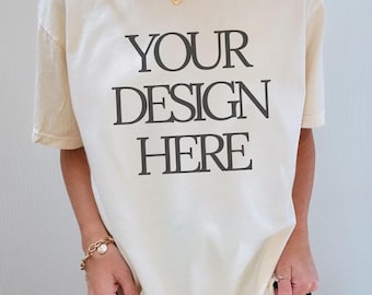 May include: A cream-colored short-sleeve t-shirt featuring the black text "YOUR DESIGN HERE" printed in a bold, serif font on the front. The shirt has a crew neckline and a relaxed fit, providing a blank canvas for custom graphic designs.