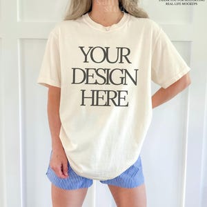 Könnte beinhalten: Cremefarbenes T-Shirt mit dem Text "YOUR DESIGN HERE" in Schwarz. Das Model trägt blau-weiß gestreifte Shorts. Das Shirt ist ein Mock-up, bereit für individuelle Designs.