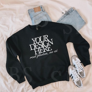 May include: A black sweatshirt with the text "YOUR DESIGN HERE real photos, no ai" in white, paired with light-wash denim jeans and white sneakers with gray accents. The items are arranged on a cream-colored surface.