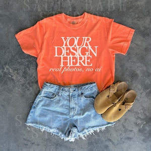 May include: An orange t-shirt with the text "YOUR DESIGN HERE real photos, no ai" is paired with light blue denim shorts and tan sandals. The shorts have a frayed hem. The sandals have a buckle closure.