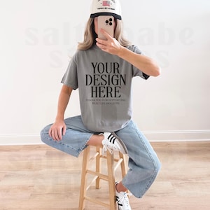 May include: A person wearing a gray t-shirt with the text "YOUR DESIGN HERE" and a white baseball cap with a black brim. The person is wearing light blue jeans and white sneakers with black stripes, sitting on a wooden stool.