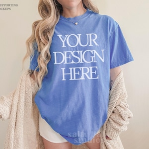 May include: A blue t-shirt with the text "YOUR DESIGN HERE" in white block letters. The person is wearing a beige cardigan and white shorts. The image includes the text "THANK YOU FOR SUPPORTING REAL LIFE MOCKUPS".
