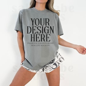 May include: A gray t-shirt with the text "YOUR DESIGN HERE" in large, bold, black letters. Below the text is "THANK YOU FOR SUPPORTING REAL LIFE MOCKUPS" in smaller black letters. The model is wearing patterned shorts.