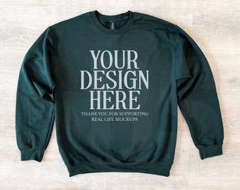 May include: A dark green long-sleeve crewneck sweatshirt featuring the light gray text YOUR DESIGN HERE centered on the chest, followed by the smaller text THANK YOU FOR SUPPORTING REAL LIFE MOCKUPS.