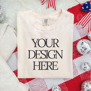 May include: Cream-colored t-shirt with the text "YOUR DESIGN HERE" in a bold, black font. The shirt is folded neatly on a red and white checkered surface, with decorative elements like disco balls and patriotic ribbons.