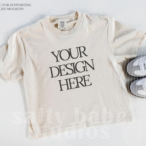 May include: Cream-colored t-shirt with the text "YOUR DESIGN HERE" in black, paired with white sneakers with black stripes. The shirt is laid flat, showcasing a mockup design. The text "THANK YOU FOR SUPPORTING REAL LIFE MOCKUPS" is at the top.