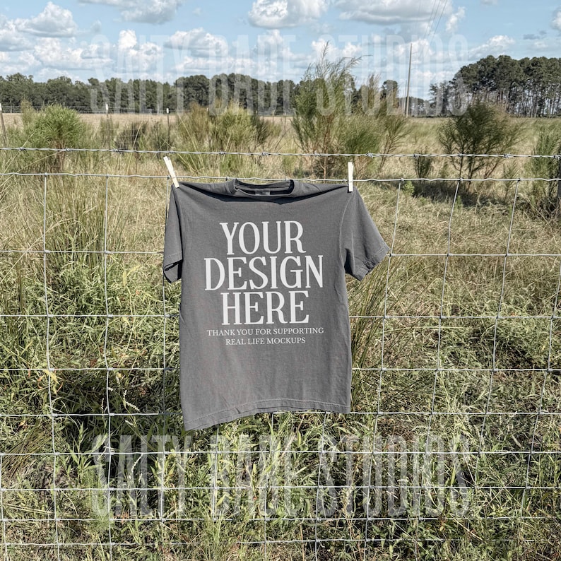 May include: A charcoal gray t-shirt hangs on a clothesline against a backdrop of a field and a cloudy sky. The shirt features the text "YOUR DESIGN HERE" in large white letters, with smaller text below that reads "THANK YOU FOR SUPPORTING REAL LIFE MOCKUPS."