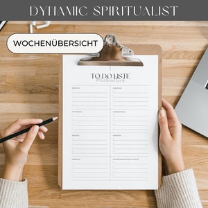 K&ouml;nnte beinhalten: Ein Holz-Klemmbrett mit einer To-Do-Liste wird von H&auml;nden gehalten, mit einem Bleistift &uuml;ber dem Papier. Ein Laptop und ein Notizbuch liegen auf dem Holztisch. Der Text "DYNAMIC SPIRITUALIST" und "WOCHEN&Uuml;BERSICHT" sind sichtbar.