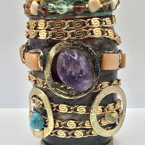 Puede incluir: Un tarro de vidrio decorativo con un borde dorado y una variedad de piedras y cuentas de colores. El tarro está decorado con cadenas y círculos dorados. El tarro es de color morado, marrón y dorado.