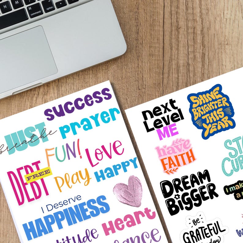 Printable 2026 Vision Board Words sheets on a wooden desk beside a laptop, featuring colorful motivational phrases like Just Breathe, Success, Shine Brighter This Year, Dream Bigger, and I Deserve Happiness. A perfect digital download for vision boards, goal setting, journaling, and New Year inspiration projects.
