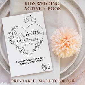 May include: A white wedding activity book with the text "KIDS WEDDING ACTIVITY BOOK" at the top. The cover features a heart-shaped floral design with the names "Mr. & Mrs. Williamson" and the date "06.07.25". The text "A happy little book for a happily ever after!" is also included.
