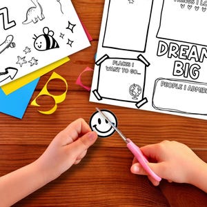 Child cutting out a smiley face from a printable Kids Vision Board 2026 craft sheet with scissors on a wooden table. Fun goal setting activity and New Year project for children featuring coloring pages, kid affirmations, and Dream Big vision board templates for creative goal planning.