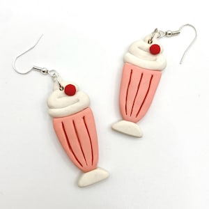 May include: Novelty earrings in the shape of pink milkshakes, topped with white whipped cream and a red cherry. The earrings have silver-coloured fish hook ear wires.