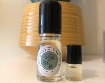 May include: Two small glass bottles with black roll-on tops. The bottle on the left has a white label with the text "ATTAR: ACTION" and a floral design. The bottle on the right is empty.