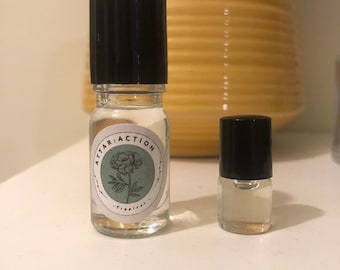 May include: Two glass bottles with black roll-on tops. The larger bottle has a white label with the text "ATTAR:ACTION" and a green floral design. The text "Tropical" is below the design.