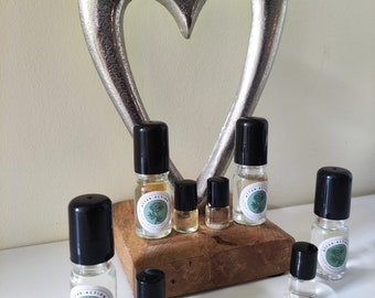 May include: A set of eight small glass bottles with black caps and labels that say "Attar Action". The bottles are arranged on a wooden surface in front of a silver heart-shaped object.