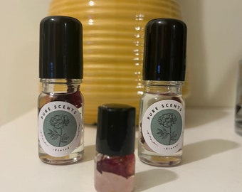 May include: Three glass bottles with black roll-on tops filled with a clear liquid and red flower petals. The bottles have a white label with the text "PURE SCENTS" and a green circle with a line drawing of a flower.