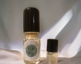 May include: Two clear glass perfume bottles with black caps. The larger bottle has a label with the text "ATTAR:ACTION" and a floral illustration. The smaller bottle is next to the larger one. The label also says "Hot Chocolate".