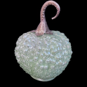 May include: A decorative glass object with a light green, textured surface and a silver-colored stem. The object has a round shape with a bubbly texture, and the stem curves upwards. The background is black.