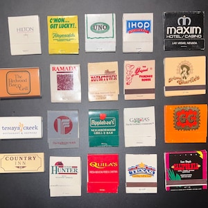 May include: A collection of vintage matchbook covers featuring various Las Vegas businesses, including hotels, restaurants, and casinos. The matchbooks are arranged in rows on a dark surface. Some of the matchbooks feature colorful graphics and logos, while others are more simple in design.