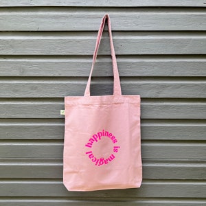 Könnte beinhalten: Eine rosa Canvas-Tragetasche mit dem Text "happiness is magical", der in einem Kreis in rosa gedruckt ist.