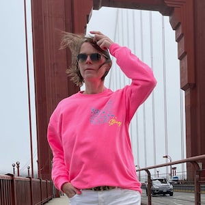 Könnte beinhalten: Eine Frau trägt ein rosafarbenes Sweatshirt mit einer blauen und gelben Grafik, auf der "Be Strong" steht. Sie steht vor einer roten Brücke.
