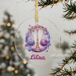 Puede incluir: Un adorno redondo de vidrio con una ilustración de acuarela del signo del zodíaco Libra. La balanza es dorada con planetas morados y azules a cada lado. El fondo es una acuarela de morado y azul con estrellas doradas. La palabra "Libra" está escrita en cursiva rosa debajo de la balanza.