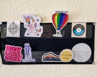 May include: A black wall organizer with clear pockets, holding various stickers. Stickers include a unicorn, a hot air balloon, and text that reads "HOT GIRL SHIT", "Pickleball is Life", and "you are the magic".