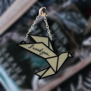 May include: A cream-colored origami bird pendant with black outlines and the text "I need you...". The pendant hangs from a chain attached to a gold-colored crescent moon and star charm. The background is blurred.