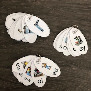 May include: Three sets of white oval-shaped flashcards with different vowel sounds and pictures. The flashcards are attached together with a metal ring. The vowel sounds include 'ew', 'oi', 'ea', 'a-e', 'ay', 'o', 'i', 'c'.