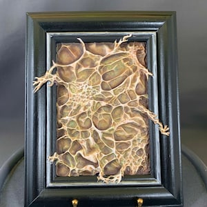 Może przedstawiać: Ramka z obrazem przedstawiającym teksturalny, organiczny, abstrakcyjny wzór w odcieniach brązu, beżu i zieleni. Obraz jest oprawiony w czarną ramę z srebrnym akcentem.