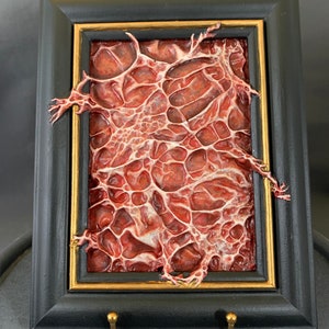 Może przedstawiać: Ramka z obrazem przedstawiającym trójwymiarowy, organiczny wzór w kolorze czerwonym i białym. Rama jest czarna z pozłacanym wykończeniem.