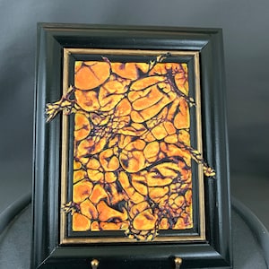 Może przedstawiać: Abstrakcyjny obraz w ramie z fakturowanym, pomarańczowo-żółtym tłem. Obraz ma czarną ramę ze złotym wewnętrznym obramowaniem.