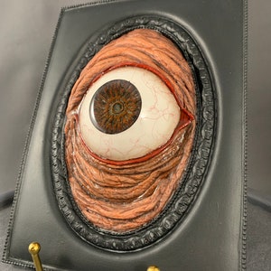 May include: A framed, realistic-looking, sculpted eyeball with a red, fleshy, wrinkled eyelid. The frame is black with a gold-colored stand.