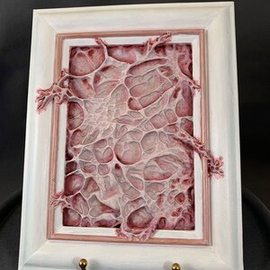 Może przedstawiać: Prostokątne dzieło sztuki w białej ramie z teksturowanym, organicznym wzorem w odcieniach czerwieni i bieli. Rama ma złoty haczyk na dole. Grafika ma unikalny, abstrakcyjny wygląd.