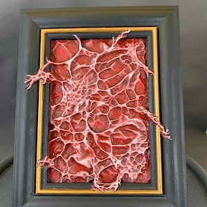 Może przedstawiać: Ramka z obrazem przedstawiającym czerwoną, fakturowaną, organiczną formę przypominającą koral lub grzyb. Rama jest czarna z pozłacanym wykończeniem.