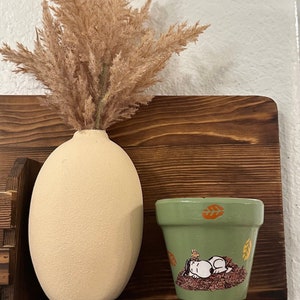 Puede incluir: Una maceta de cerámica verde con una ilustración pintada a mano de Snoopy durmiendo en un montón de hojas de otoño. La maceta está sentada en un estante de madera junto a un jarrón beige con hierba de la pampa seca.