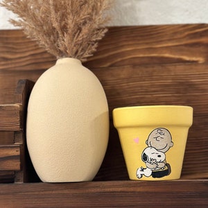May include: Yellow ceramic flower pot featuring a hand-painted design of Snoopy and Charlie Brown embracing. The pot is displayed on a wooden shelf with a vase of dried pampas grass.