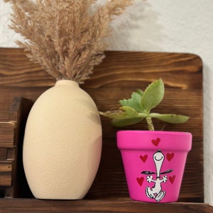 May include: A pink terracotta planter with a hand-painted design of Snoopy holding hearts. The planter is filled with a small green succulent plant.
