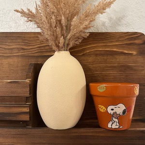 Puede incluir: Un jarrón de cerámica beige con hierba de la pampa seca y una pequeña maceta de terracota naranja con un diseño pintado de Snoopy y hojas de otoño.