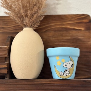 Puede incluir: Una maceta de cerámica azul con un diseño pintado a mano de Snoopy durmiendo en una luna creciente con estrellas.