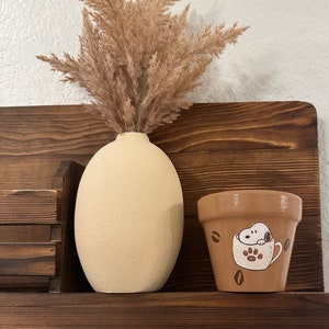 Puede incluir: Un jarrón beige de forma ovalada contiene hierba ornamental seca de color marrón claro. A la derecha, una pequeña maceta marrón presenta una ilustración de Snoopy dentro de una taza de café, con acentos de granos de café. Los objetos descansan sobre una estantería de madera marrón oscuro.