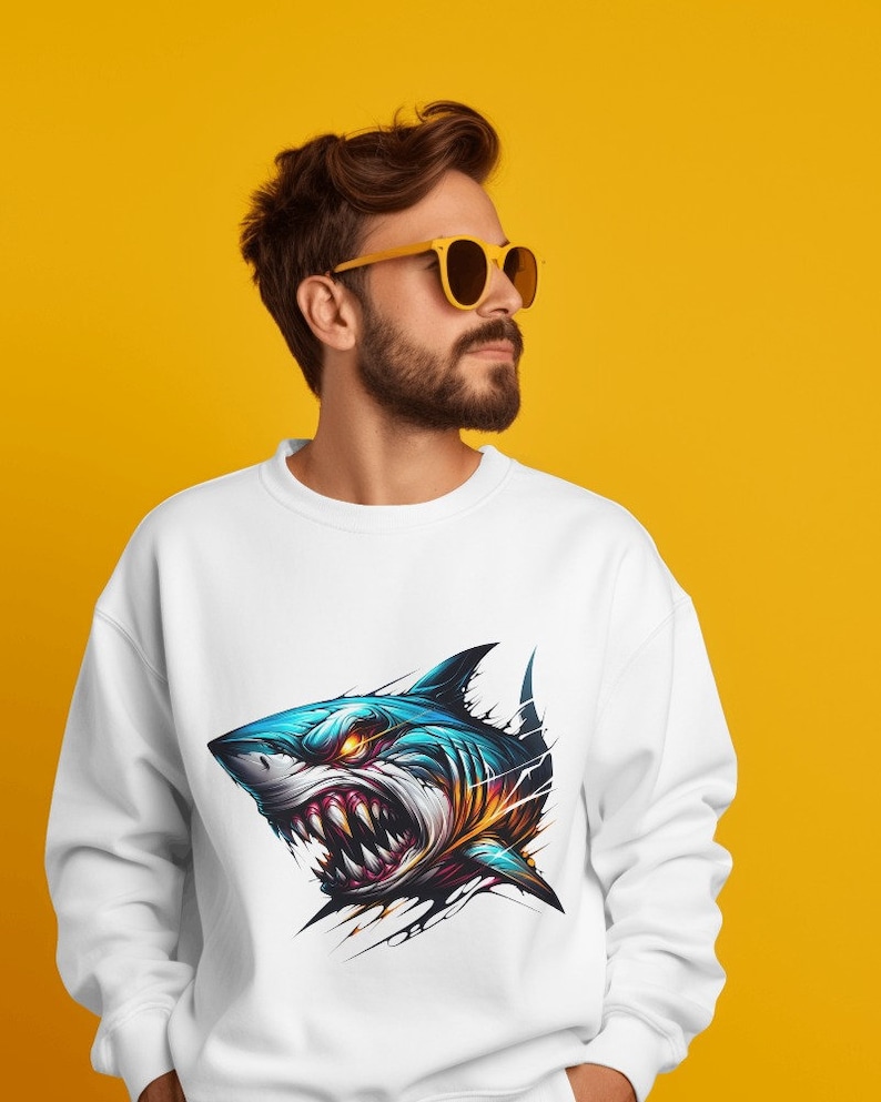 May include: A white sweatshirt with a graphic print of a blue and yellow shark with sharp teeth and angry eyes. The shark is depicted as if it is breaking through the fabric of the shirt.