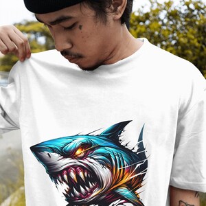 May include: White t-shirt with a graphic of a blue and orange shark with sharp teeth and glowing eyes. The shark is depicted as breaking through the fabric of the shirt.