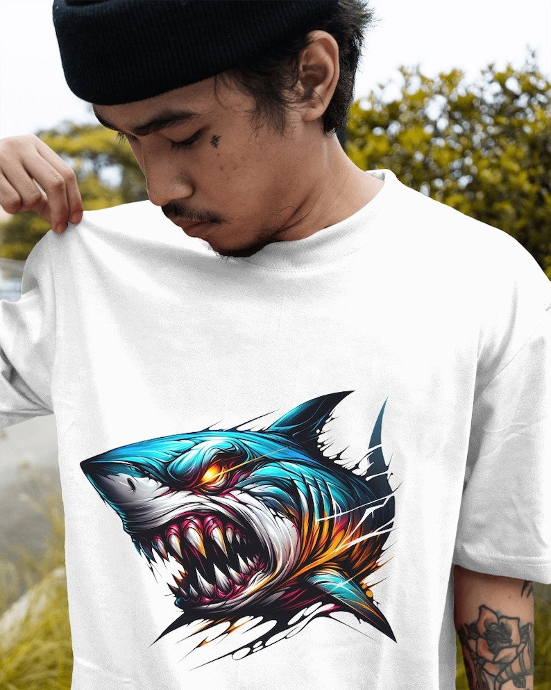 May include: White t-shirt with a graphic of a blue and orange shark with sharp teeth and glowing eyes. The shark is depicted as breaking through the fabric of the shirt.