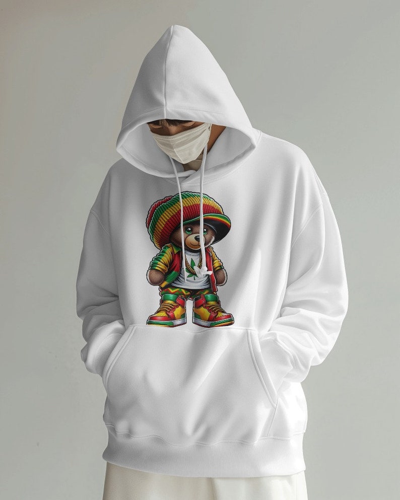 Puede incluir: Sudadera con capucha blanca con un estampado gr&aacute;fico de un oso de dibujos animados que lleva un gorro rojo, amarillo y verde, una chaqueta roja y pantalones amarillos. El oso sostiene una hoja verde de marihuana.